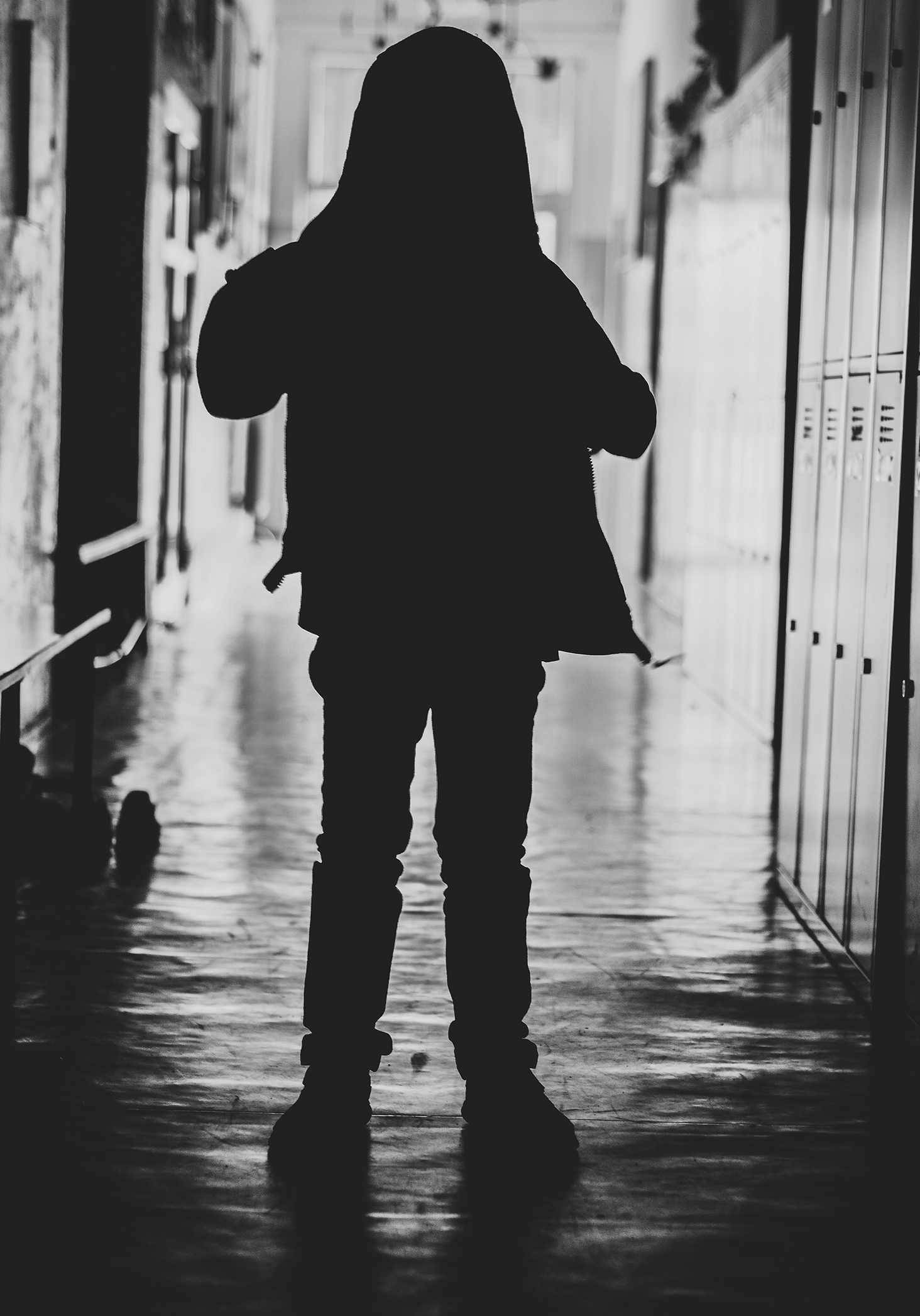 A young child standing alone in a school hallway, wearing a sweater and cast completely in a black silhouette